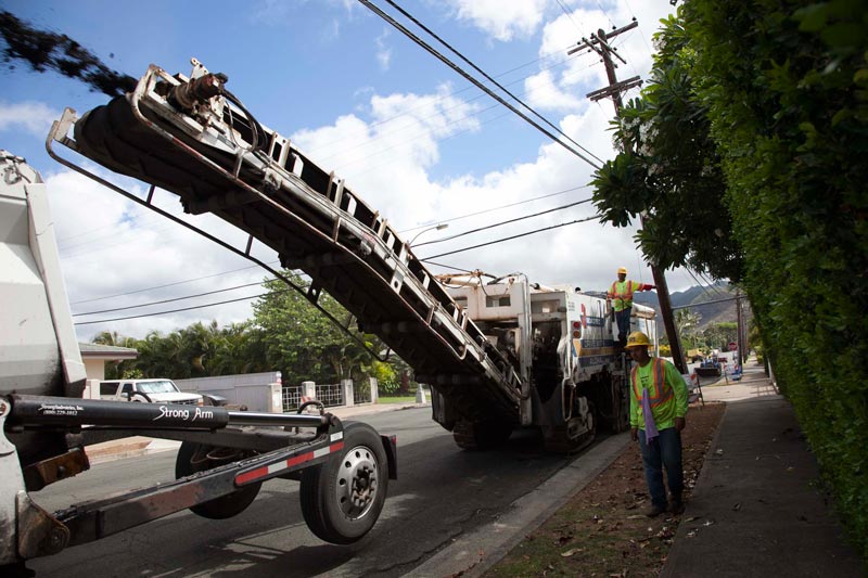 A milling machine transfers removed pavement to a truck on a neighborhood street.