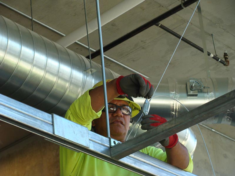 A worker installs metal framing beneath overhead ductwork.